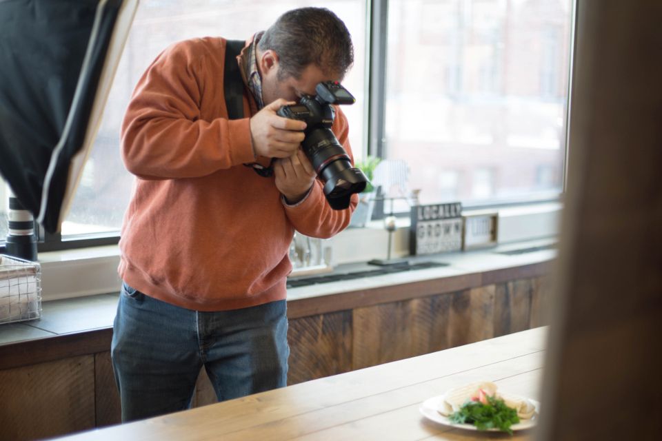 A photographer in an orange sweater shoots a plated dish on a restaurant counter by a window.