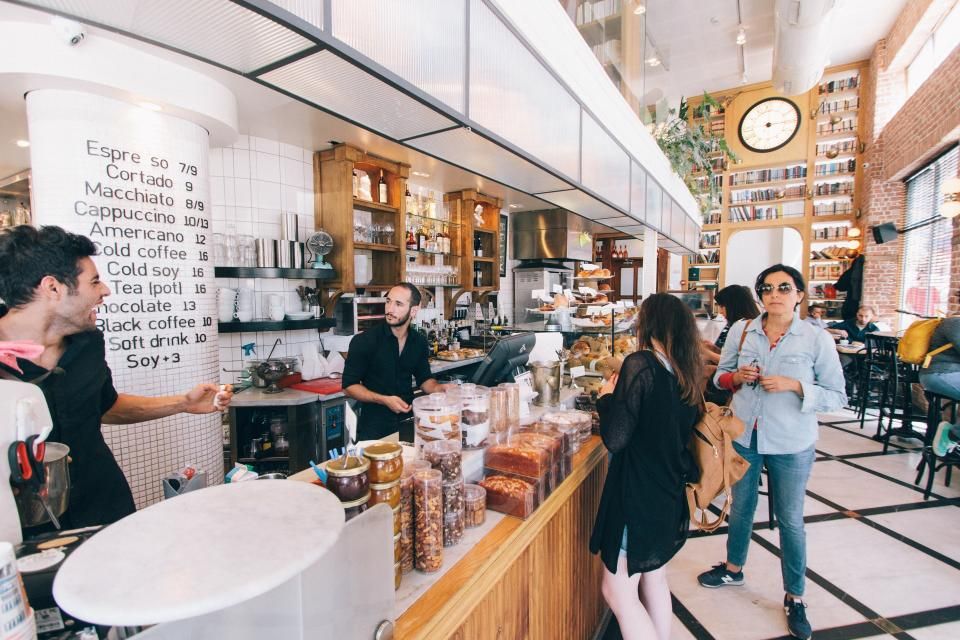 Staff behind a café counter serve customers, with a handwritten drinks menu on a white pillar.