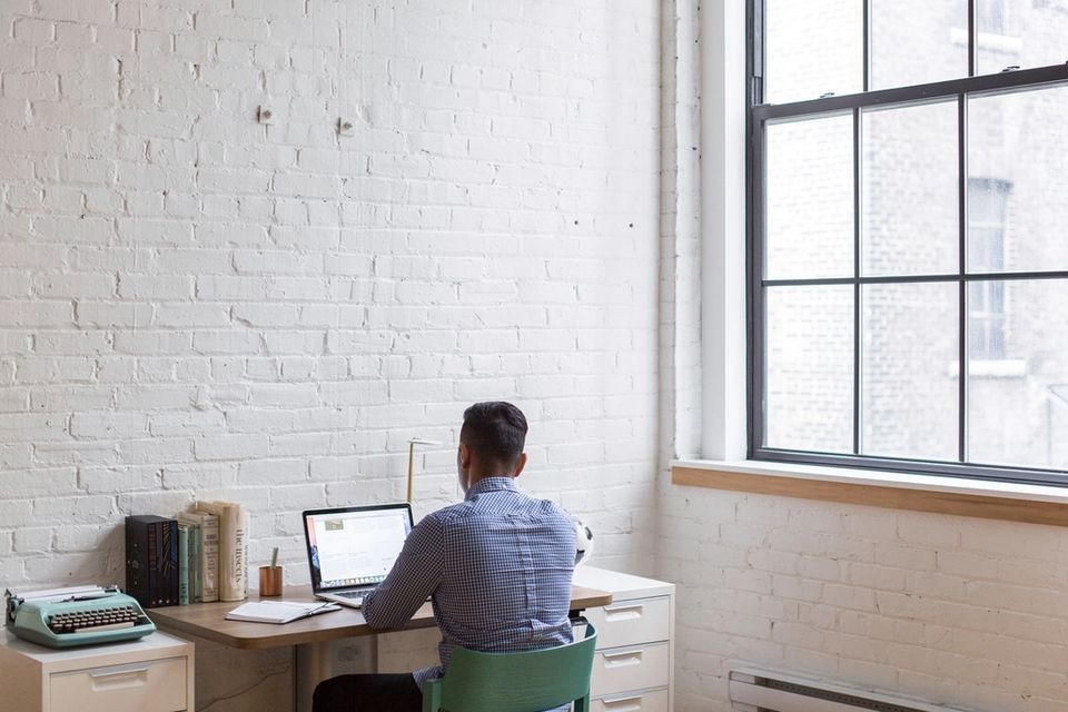 A man works on a laptop at a small desk against a white brick wall, next to a tall window.